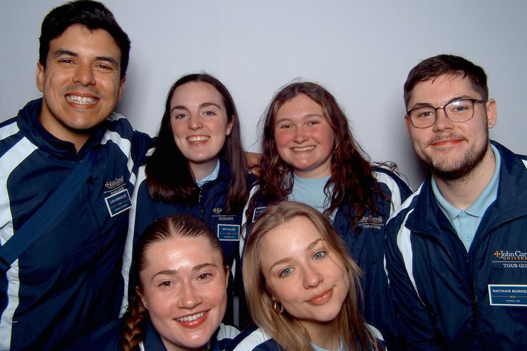 Six young adults wearing matching John Carroll University tour guide jackets smile closely together against a plain.