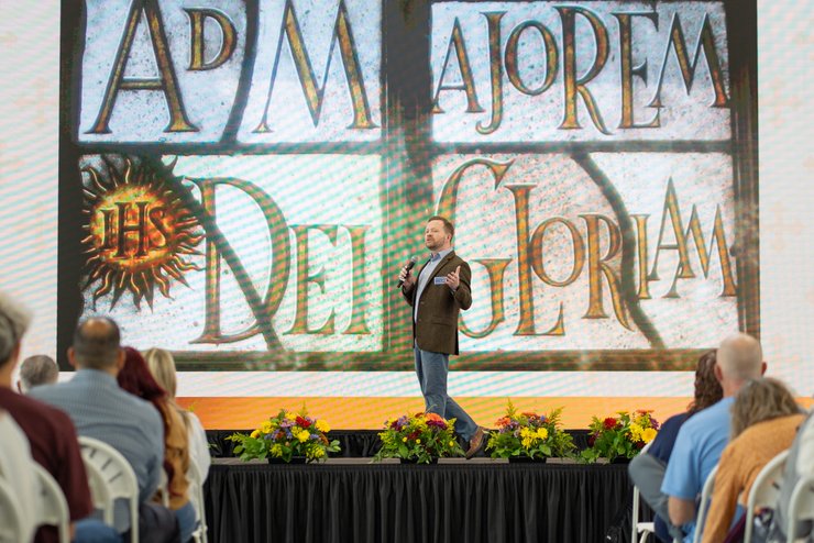 A man stands on a stage speaking into a microphone in front of an audience and a large screen with stylized text.