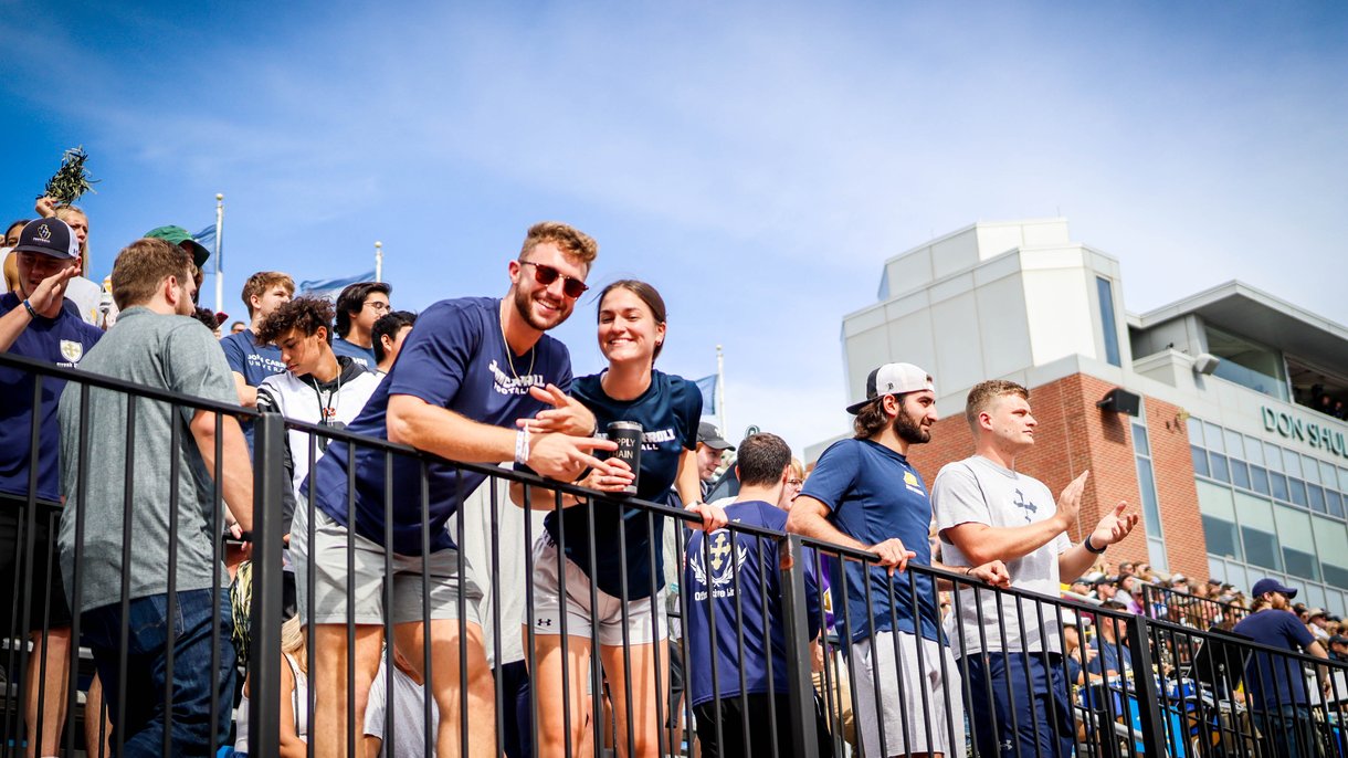A group of young people stand behind a black railing in a stadium, some smiling and clapping under a blue sky.