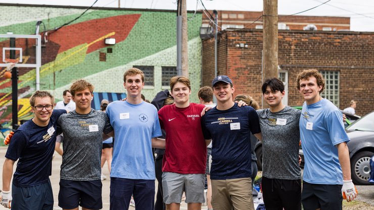 Seven young men stand arm in arm smiling outdoors near a basketball hoop and a colorful mural on a brick wall.