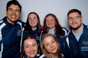 Six young adults wearing matching John Carroll University tour guide jackets smile closely together against a plain.