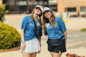 Two young women wearing blue John Carroll University shirts and white caps smile and lean toward the camera outdoors.