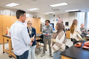 A group of six people stand and talk in a classroom with anatomical models and posters on the walls.