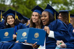 Three graduates in blue caps and gowns smile and hold up their diploma covers outdoors.