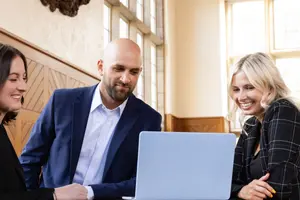 Three people in business attire smile and look at a laptop in a room with large windows and wood paneling.