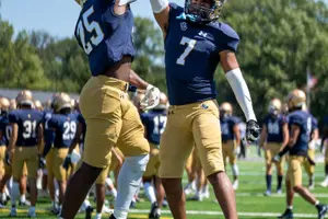 Two football players in navy and gold uniforms jump and high-five on a green field under a clear blue sky.