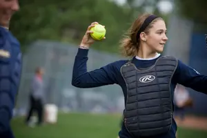 A softball player wearing a chest protector prepares to throw a yellow softball during practice on a grassy field.