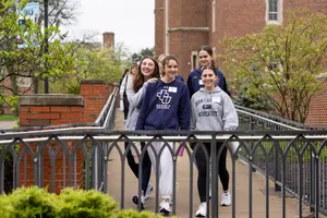 Four young women walk together on a paved path beside a brick building, smiling and wearing casual sweatshirts.