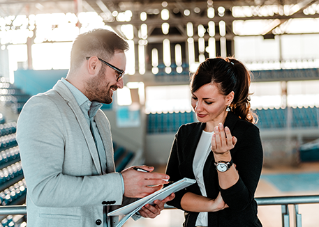 Picture of male (left) and female (right) coaches in gymnasium discussing information in notebook