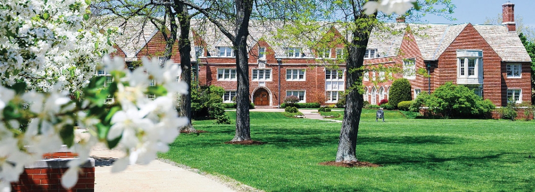 Rodman Hall across a grassy field with large tall trees and bushes blooming with vibrant white flowers.