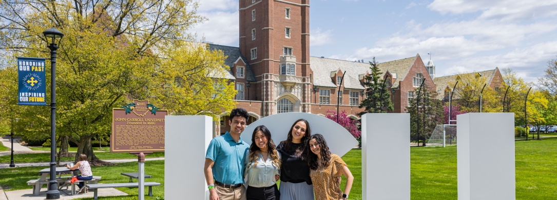 Four students posing in front of the JCU clock tower with white bold letters spelling JCU