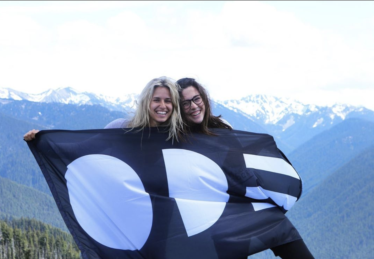 Spear and O'Connell smile on a mountaintop holding an Ode flag