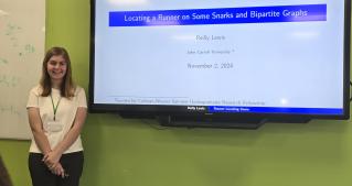 JCU mathematics major Reilly Lewis stands next to classroom display screen with title slide of her mathematics research presentation